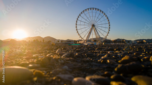 aerial drone shot of a Sunrise over Marseille’s waterfront with Ferris wheel, calm shoreline, rocky jetty, and golden mountains creating a serene coastal cityscape