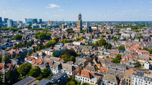 Aerial drone view of Utrecht, Netherlands featuring its historic church tower, red‑roofed buildings, modern skyline, and sunny urban landscape blending old and new architecture.