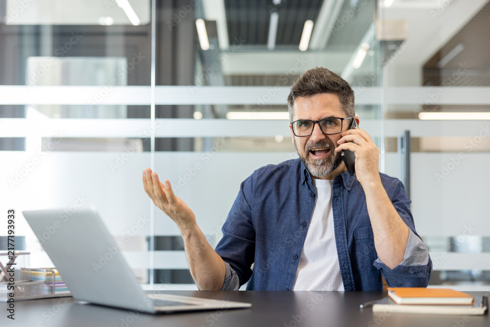 Obraz premium Mature businessman experiencing stress and frustration, shouting into his mobile phone while working at his office desk, indicating conflict and professional burnout