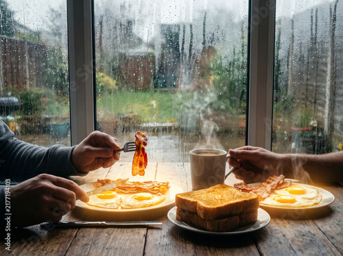 Cozy breakfast for two by rainy window with fried eggs bacon and steaming coffee.