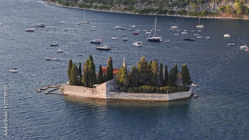 aerial view of st george island with its tall cypress trees and stone monastery wall on deep blue water, with dozens of anchored boats along the opposite shore, bay of kotor, montenegro