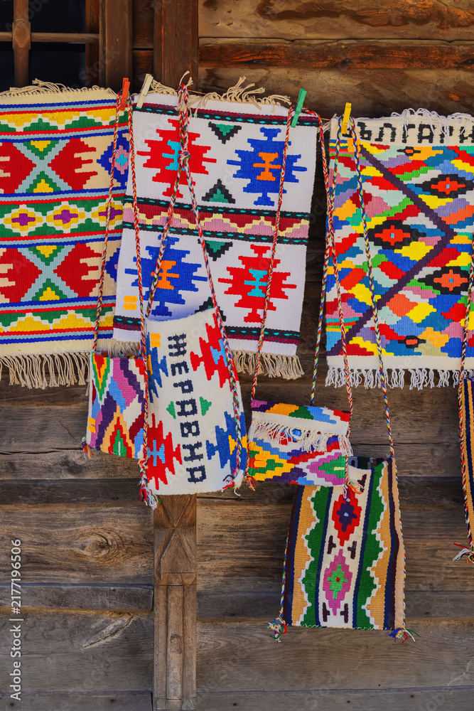 Obraz premium Traditional Bulgarian woven rugs and handmade textile bags. Colorful Bulgarian woven rugs and handmade textile bags displayed on rustic wooden wall. Authentic folk craft, vibrant ethnic ornament