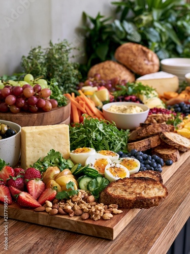 Wooden platter filled with a variety of fresh fruits and vegetables. on the left side of the platter, there is a wooden bowl filled with grapes, olives, and a small bowl of cheese.