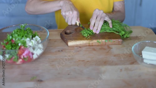 Woman chopping fresh dill on wooden cutting board while preparing healthy vegetable salad with feta cheese in kitchen