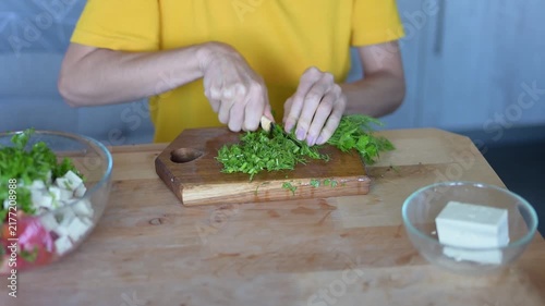 Woman chopping fresh dill on wooden cutting board while preparing healthy vegetable salad with feta cheese in kitchen