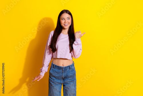 Young woman smiling and pointing to the side on a bright yellow background wearing a cropped pink top and denim jeans