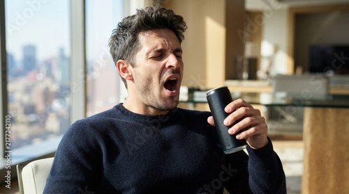 Tired man yawning widely while holding a black energy drink can. Sleepy male in a blue sweater sitting by a window with city views