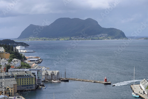 Scenic view of Alesund coastal town harbor with a small red lighthouse and mountains in Norway. Beautiful norwegian cityscape of Alesund port with boats and Sukkertoppen mountain in the background.