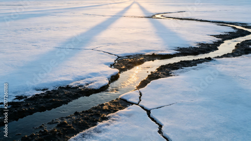 Melting crack on frozen snow field, winding stream with golden sunrise light