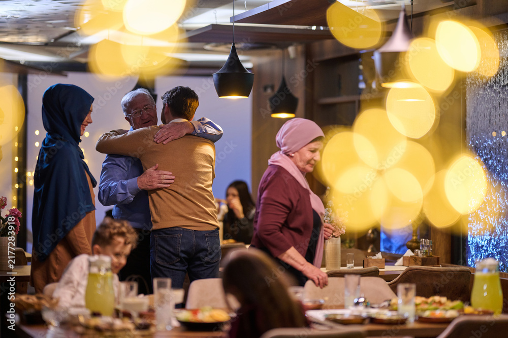 Naklejka premium Multi-Generational Muslim Family Hugging at Festive Dinner Reunion With Warm Lights and Food