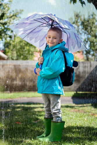 Obraz na płótnie Happy boy in a blue raincoat holds an umbrella on a rainy day, wearing boots and