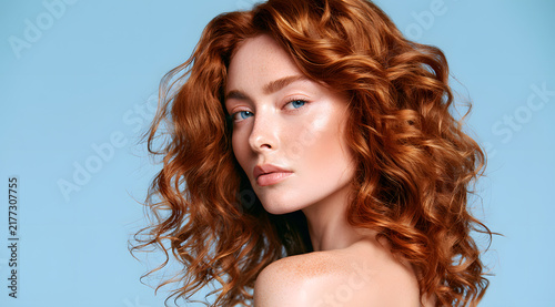 A side-view photo of a young fair-haired woman, whose hair is voluminous and wavy, with freckles and blue eyes. The skin tone is fair, and she stands against a light blue backdrop in a studio setting.