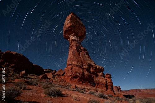 Star trails around a balanced rock formation in a desert landscape at night