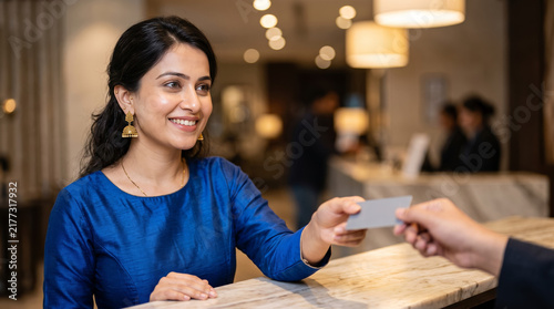 A cheerful Indian woman wearing a vivid blue top smiles warmly as she is handed a key card at a well-lit hotel reception counter, featuring elegant contemporary decor and soft ambient lighting.