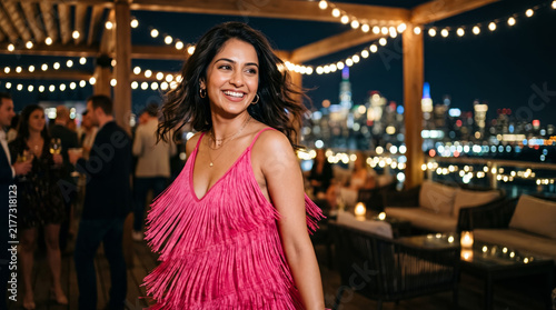 A stylish woman in a bold magenta fringe dress smiles radiantly at an upscale rooftop evening event. String lights and a stunning illuminated city skyline glow beautifully in the festive background.