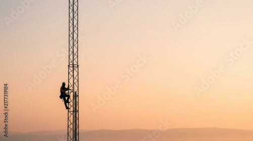 Technician climbing tower at sunset with safety gear and helmet, working on communication or electrical structure