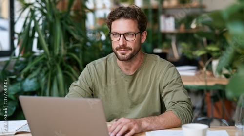A man with a beard and glasses wearing a green shirt sits at a desk with a laptop in front of him. He is surrounded by many green plants.