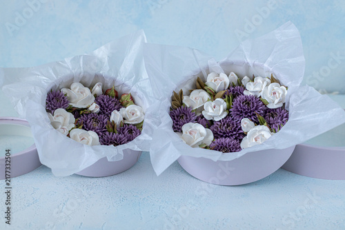 Floristry from Zephyr. Floral arrangement in round purple boxes featuring white roses and purple asters with decorative tissue paper on a light blue background