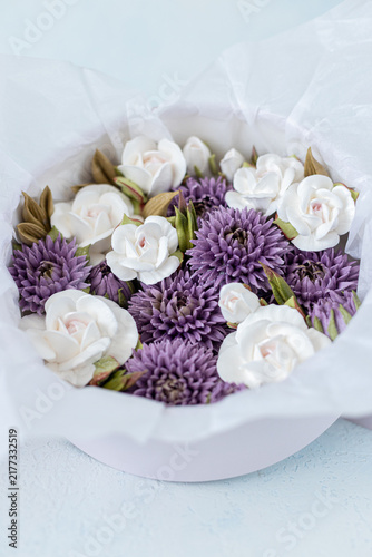 Floristry from Zephyr. Arrangement of white roses and purple chrysanthemums in a round box with tissue paper, set against a light blue background