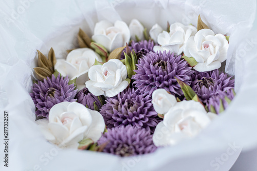 Floristry from Zephyr. White and purple artificial flowers arranged in a round bouquet with green leaves, wrapped in white tissue paper for decorative purposes