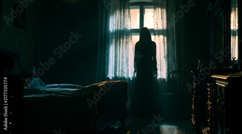 By the window in the dark bedroom, a haunting silhouette of a woman stands. Moonlight filters through the lace curtains, casting eerie shadows. An apparition drifts through the old room, with the bed 