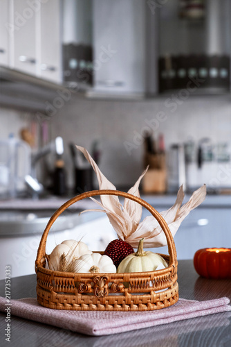 Cozy autumn still life with white mini pumpkins, pomegranate and dry corn husks in a wicker basket on a kitchen table with a burning pumpkin shaped candle. Soft blurred interior background.