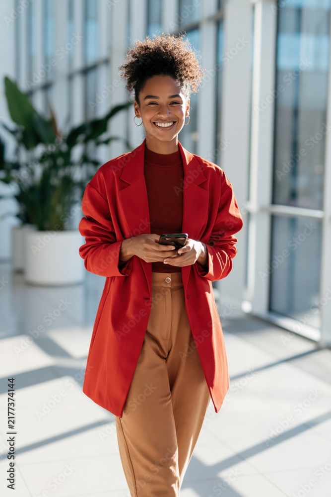 Obraz premium Happy professional woman in red coat walking with smartphone in sunlit office corridor for business banner and background design