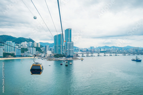 Cable car gondolas cross above the sea with Busan high-rise buildings skyline, bridge and mountains, South Korea. Elevated transport system, tourism infrastructure, coastal mobility, engineering