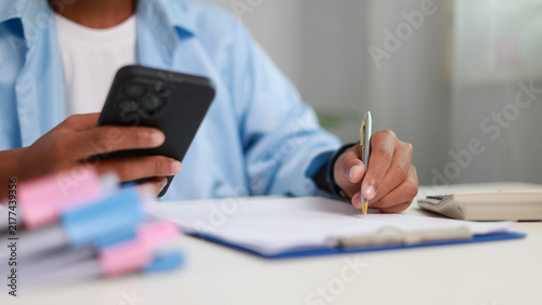 Close-up of a businessperson holding a smartphone and writing on a document with pen on desk with calculator. Multitasking office work concept.