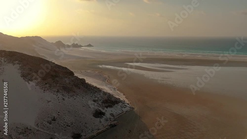 Socotra island, Aerial view of massive white sand dunes meeting the ocean under a golden sunset. Rocky cliffs mountain line the coastal landscape as bright sunlight reflects on the water. Drone flight