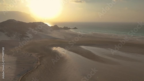 Socotra island, Aerial view of massive white sand dunes meeting the ocean under a golden sunset. Rocky cliffs mountain line the coastal landscape as bright sunlight reflects on the water. Drone flight