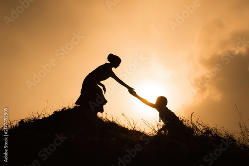 Silhouette of two people helping each other climb a steep grassy hill against a bright golden sunset with silhouette climbing teamwork cooperation adventure together connection