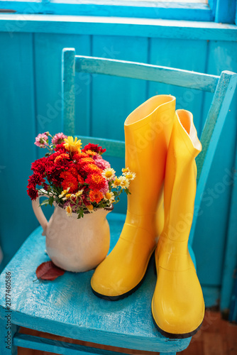 On an old blue wooden chair on an old country veranda, next to a pitcher filled with colorful autumn chrysanthemums, sits a pair of bright yellow rubber boots.