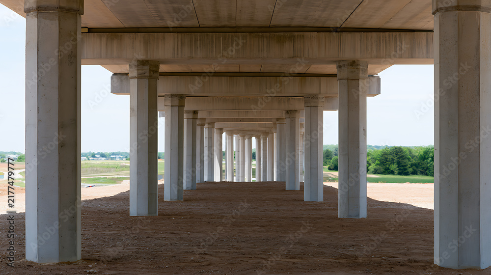 Naklejka premium Perspective view underneath a modern concrete highway overpass bridge with massive support pillars and structural framework in daylight