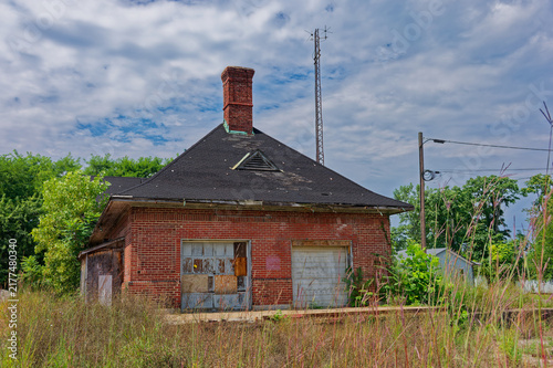 Foto An abandoned Pennsylvania Railroad red brick train station with boarded windows, an overgrown platform and adjacent railroad tracks beneath a bright blue sky