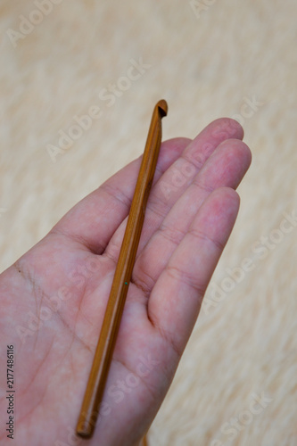Woman hand holding a single wooden crochet hook close-up against a soft beige fur background. Crochet tool, needlework supplies, craft equipment, and cozy hobby concept