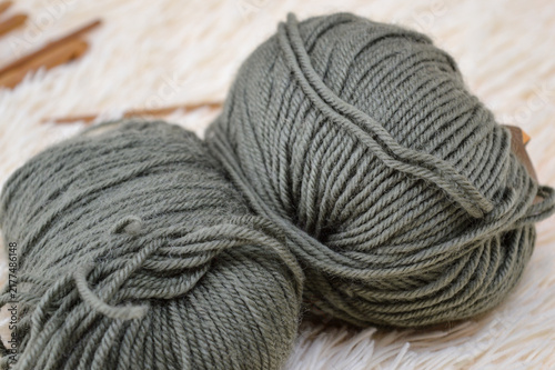 Balls of olive green wool yarn resting on a soft white fluffy fur surface with wooden crochet hooks blurred in the background. Crocheting supplies, craft materials, needlework tools, and cozy hobby co