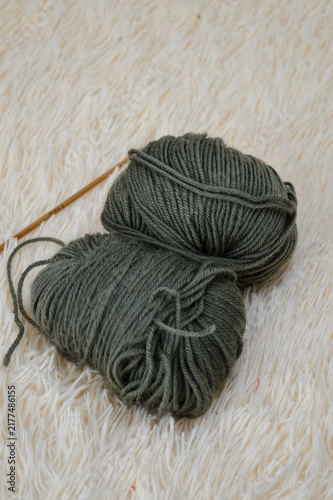 Balls of olive green wool yarn resting on a soft white fluffy fur surface with wooden crochet hooks blurred in the background. Crocheting supplies, craft materials, needlework tools, and cozy hobby co