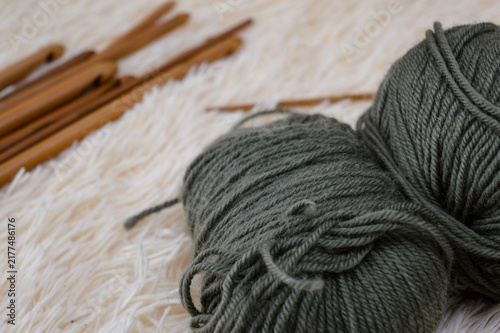 Balls of olive green wool yarn resting on a soft white fluffy fur surface with wooden crochet hooks blurred in the background. Crocheting supplies, craft materials, needlework tools, and cozy hobby co