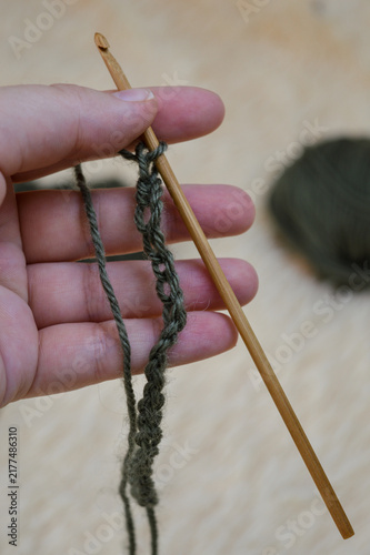 Woman hand holding a wooden crochet hook working on a green wool yarn chain close-up. Crocheting process, handmade yarn craft, needlework project, and cozy hobby concept