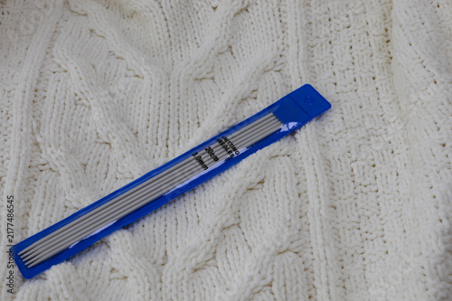 Set of double-pointed metal knitting needles in a blue plastic package resting on a white knitted woolen sweater close-up. Knitting supplies, needlework equipment, craft tools, and cozy hobby concept