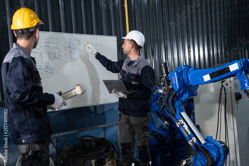 Tech specialist explaining robot motion diagram on whiteboard while colleague holds control pendant near blue mechanical arm unit