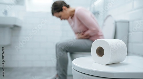 Woman suffering from stomach pain sitting on toilet with toilet paper roll in foreground representing digestive issues with experiencing stomach while sitting close up gastrointestinal cleanliness