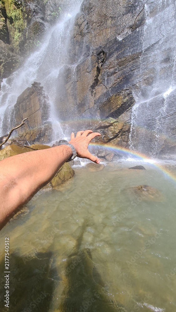 Obraz premium A person's hand reaching out to touch a beautiful rainbow created by a cascading waterfall in a natural landscape