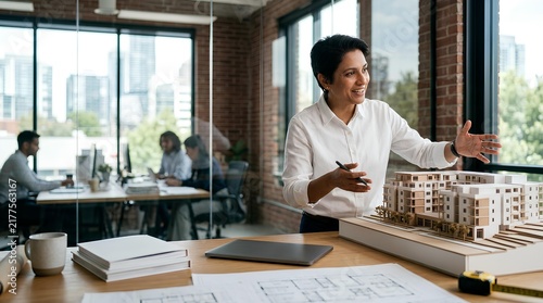 Professional female architect presenting a 3D architectural model of a residential building in a modern sunlit office with blueprints, city background, and office colleagues.