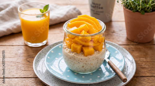 Overnight oats or yogurt parfait layered with mango chunks served on a rustic table with fresh orange juice and a green plant.