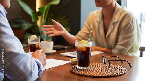 Business colleagues discussing work at a wooden table with iced coffee, documents, and eyeglasses in a bright office