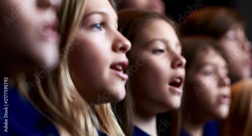 Focused young girls with open mouths singing in a school choir during a performance or rehearsal. A beautiful blonde girl with blue eyes is in the foreground, emotionally performing her part