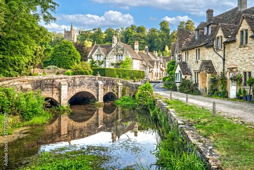 Idyllische Ansicht des historischen Dorfes Castle Combe in England mit einer alten Steinbrücke über den Fluss By Brook, traditionellen Steinhäusern und üppiger grüner Landschaft.