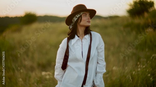 Stylish woman wearing a brown hat and white shirt walking through a grassy field at sunset